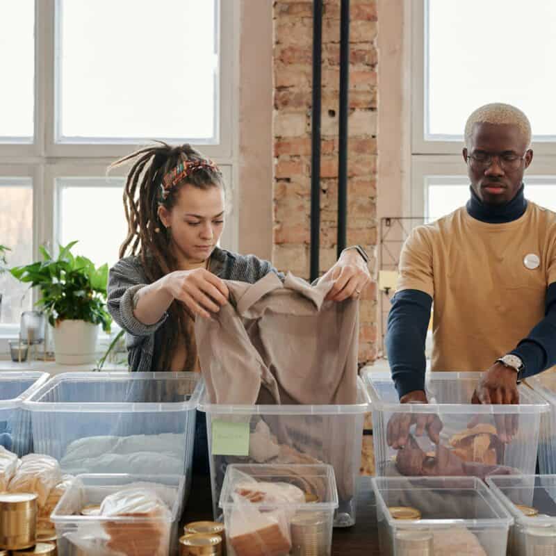 Two volunteers organize clothing and food donations indoors during daylight.